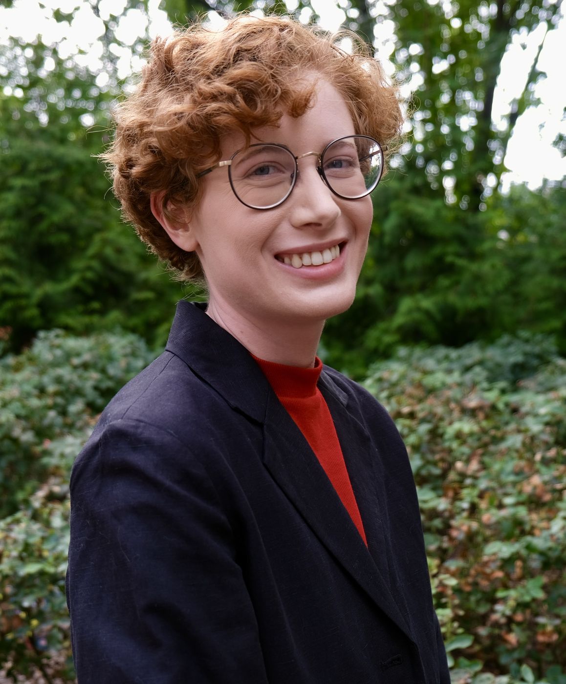 Portrait: curly copper hair, round glasses, dark blazer, smiling in front of greenery