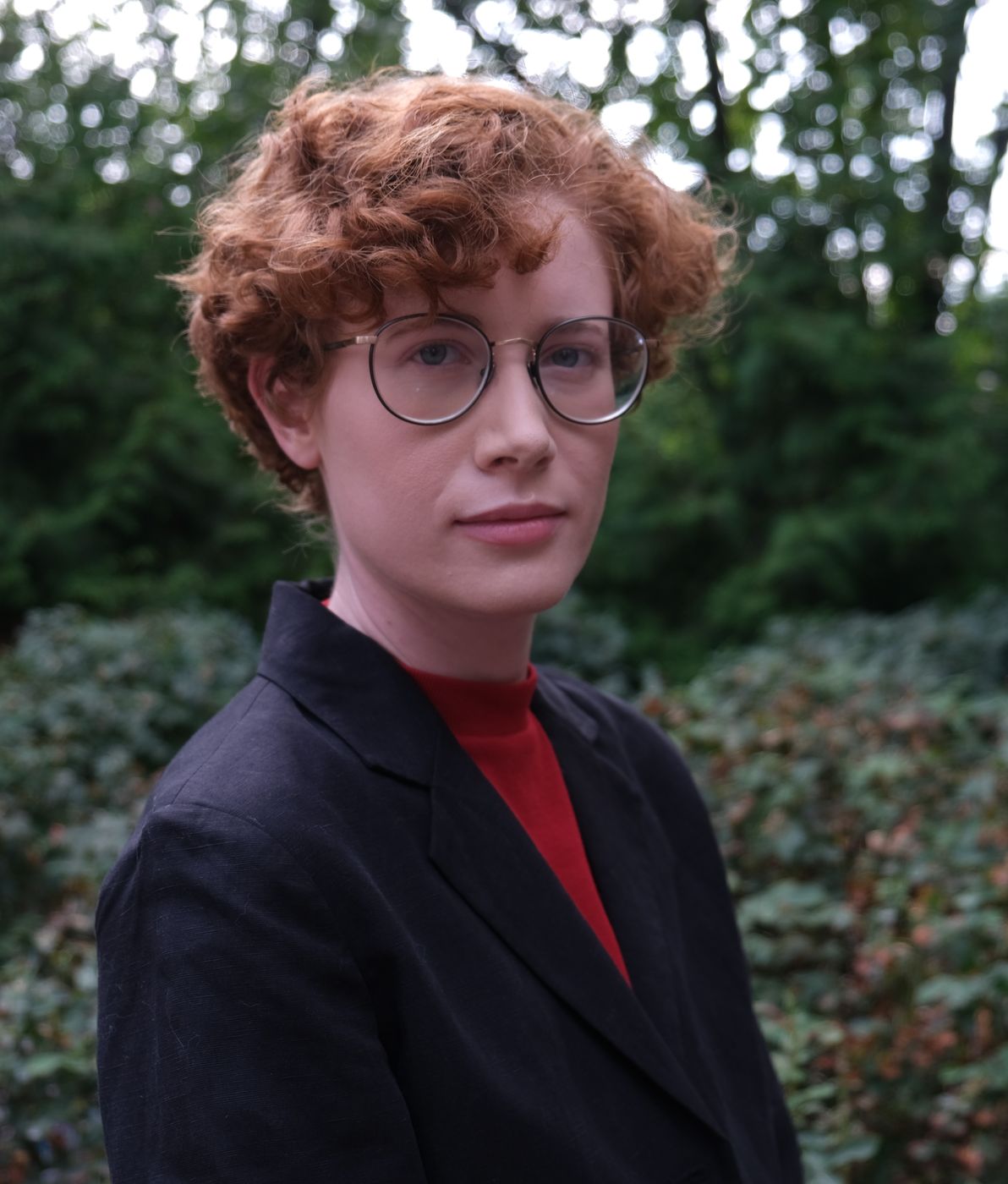 Portrait: curly copper hair, round glasses, dark blazer, in front of evergreen foliage